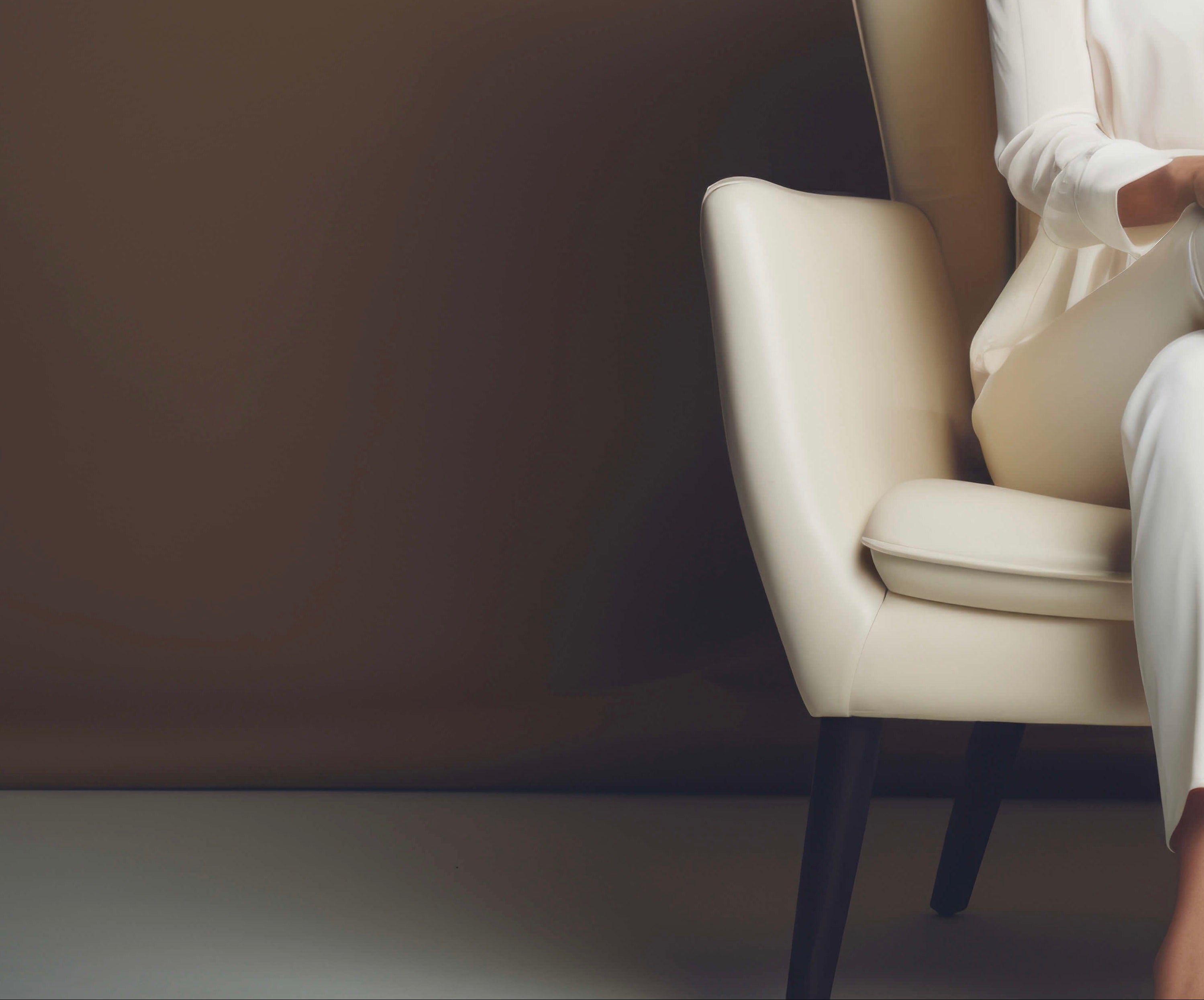 Woman in a white outfit sitting on a cream-colored chair against a neutral background