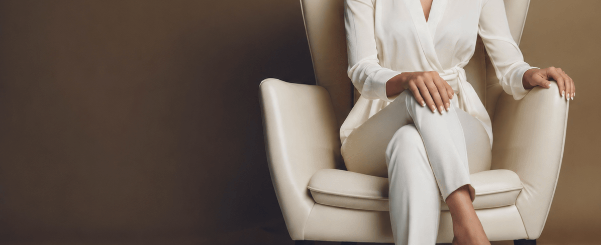 Woman in a white outfit sitting on a cream chair against a brown background